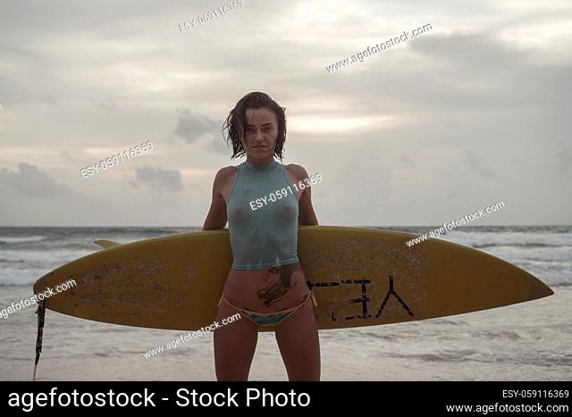 Brunette beautiful woman wearing wet top and bikini bottom standing on the beach with yellow surfboard during early evening over sea and sky background