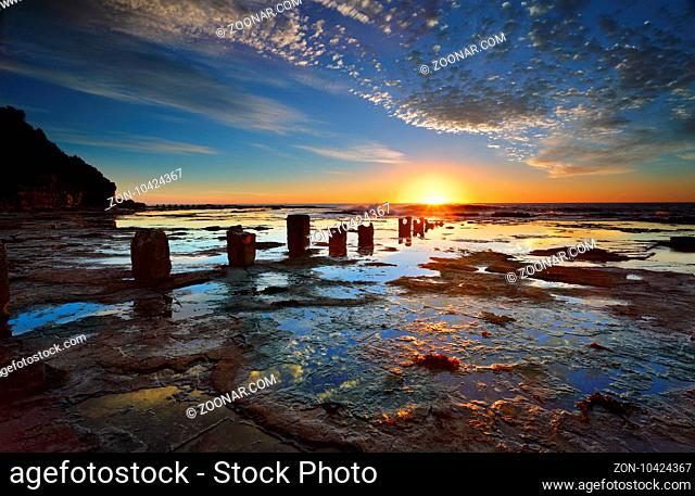 The golden sun rises on the horizon its first rays of light streaking across the large wet rockshelf and highlighting ithe clouds The surface textures of the...
