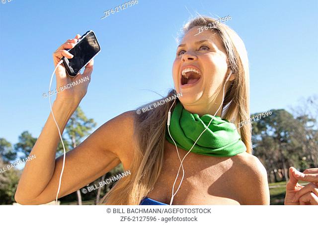 young attractive 50 year old woman listening to music on cell phone smiling outdoors still beautiful even in middle age with sunlight in her hair and great...