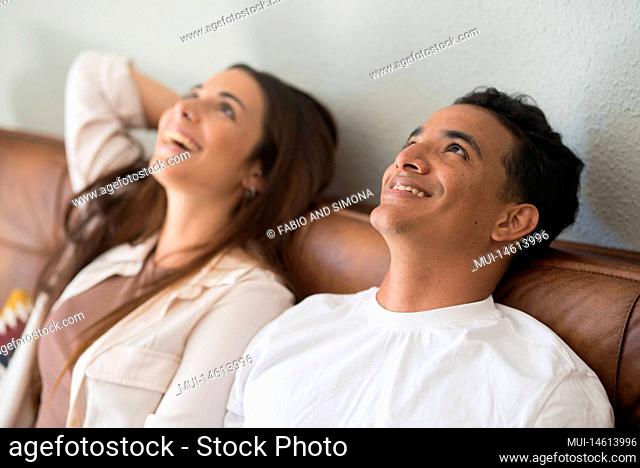 Young couple dreaming together at home sitting on the sofa and looking up with a smile. People living in a new house after moving and mortgage banking loan