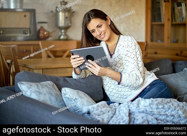 beautiful young smiling woman in white sweater sitting with a tablet on a gray sofa