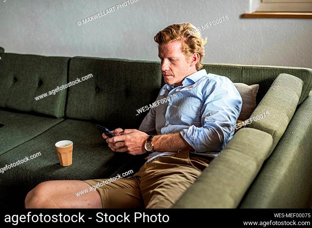 Mid adult man text messaging on smart phone while lying on couch in living room