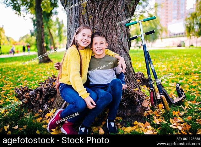 Friends kids posing happily sitting under a tree in an autumn park next to kick scooters. Happy children. Eco transport. Outdoor activities twins