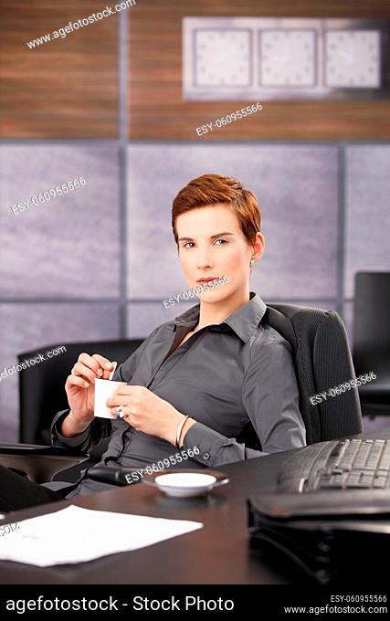 Confident trendy businesswoman drinking coffee at desk, looking at camera