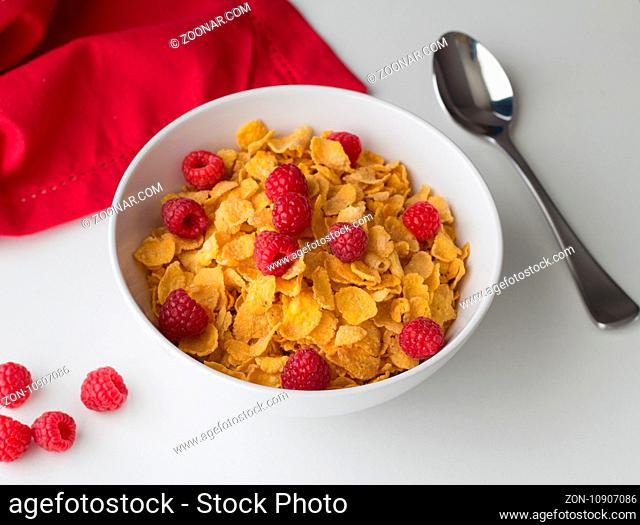 Cornflakes breakfast cereal with raspberries in bowl on white table with selective focus