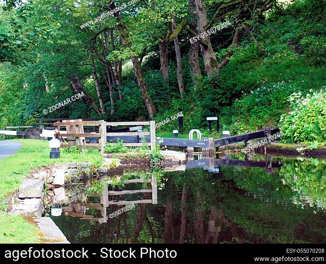 old wooden lock gates with trees and flowers reflected in the water with mooring posts and fences on the rochdale canal at rawden mill near erringden near...
