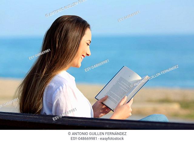 Happy woman reading a paper book sitting on a bench on the beach