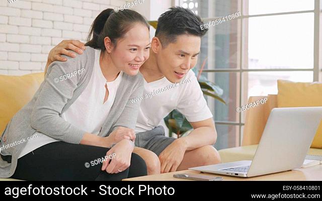 Happy Asian family couple husband and wife laughing sitting on sofa using laptop computer webcam technology talking making online social distance video call to...