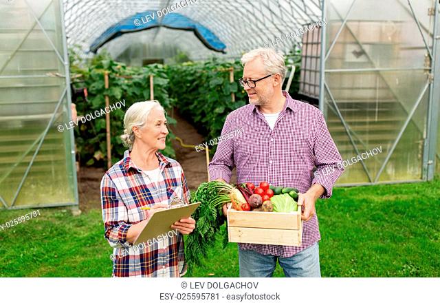 farming, gardening, harvesting and people concept - senior couple with box of vegetables and clipboard at farm greenhouse