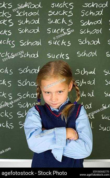 Little, angry girl standing in front of blackboard with crossed arms. Looking at camera. Front view