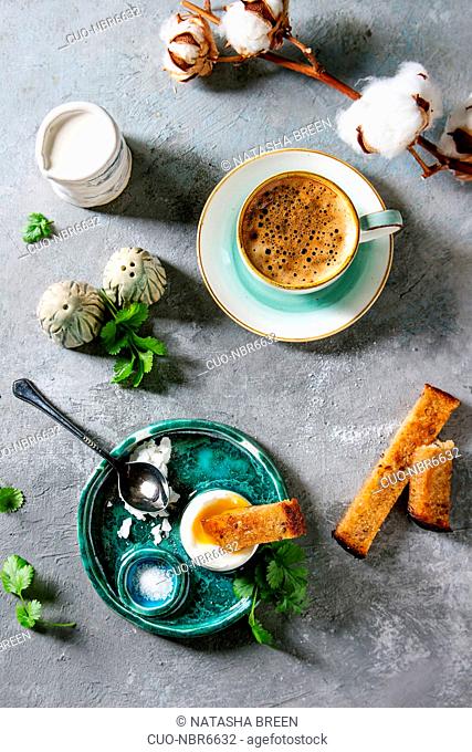 Breakfast with cup of coffee and soft boiled egg, served in green ceramic egg cup with salt, pepper and toasted bread, jug of cream and cotton flowers over grey...
