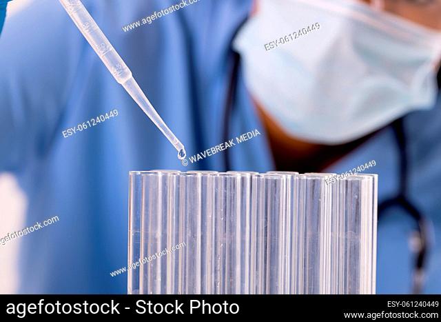 Midsection of african american mid adult female doctor wearing mask pipetting liquid in test tubes