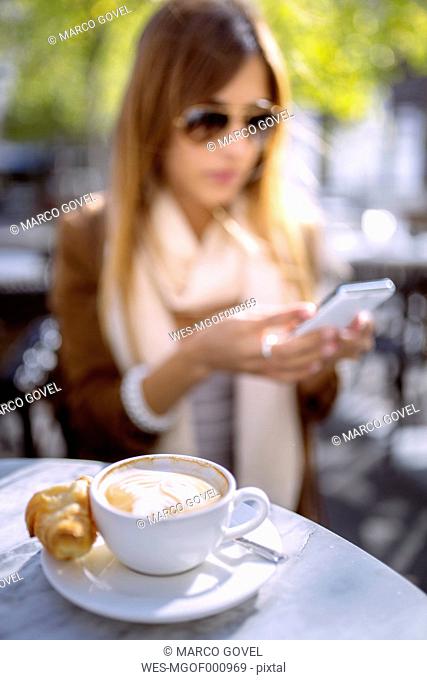 Spain, Gijon, Cup of cappucino, young woman in background using smart phone