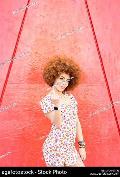 Young woman gesturing obscene hand sign while standing in front of red wall
