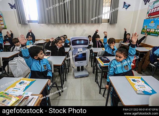 02 December 2021, Palestinian Territories, Gaza City: A home-made talking educational robot stands in the midst of Palestinian students raising their hands to...