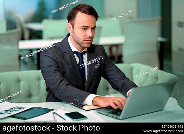 Freelancer sitting in front laptop in bright coworking space. Handsome man in business suit working on laptop, freelancer job in progress