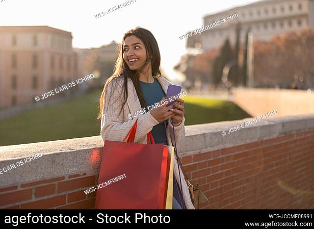 Thoughtful young woman with smart phone standing by wall
