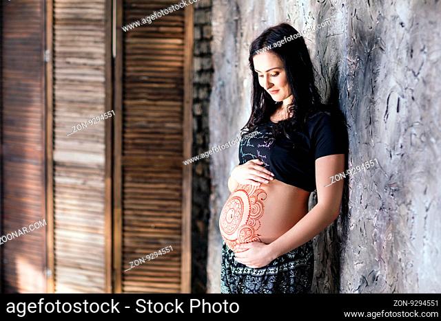 Pregnant young woman with mehendi tattoo on her belly stand near gray wall