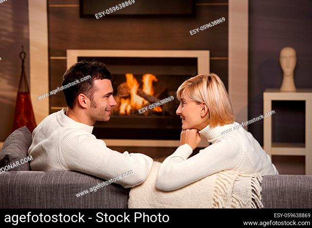 Young romantic couple sitting on sofa in front of fireplace at home, looking at each other, smiling