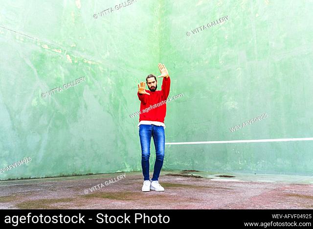 Portrait of serious young man wearing red sweatshirt raising hands in front of green wall