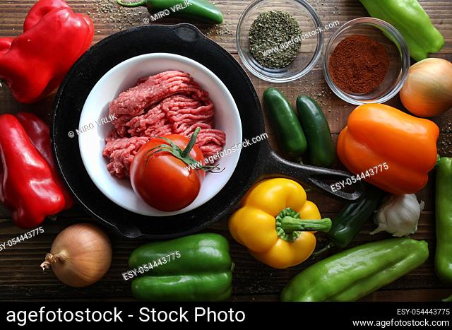 Raw ground beef inside skillet with peppers and other cooking ingredience around with wooden background top view