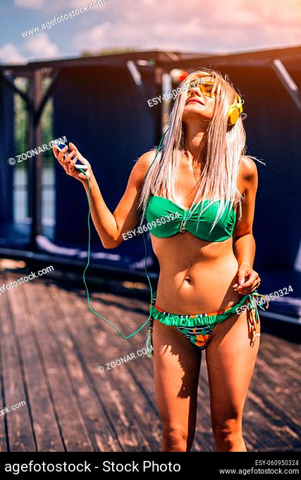 Young Woman Is Listening To Music With Yellow Headphones While Standing Near The Pool.She Is Holding Her Phone And Looking Up