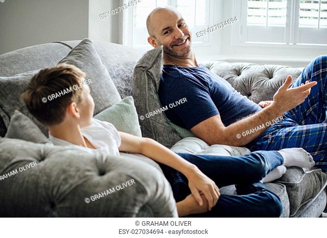 Father and son sitting on the sofa in their home together in teh morning. They are talking and laughing about something they are watching on television