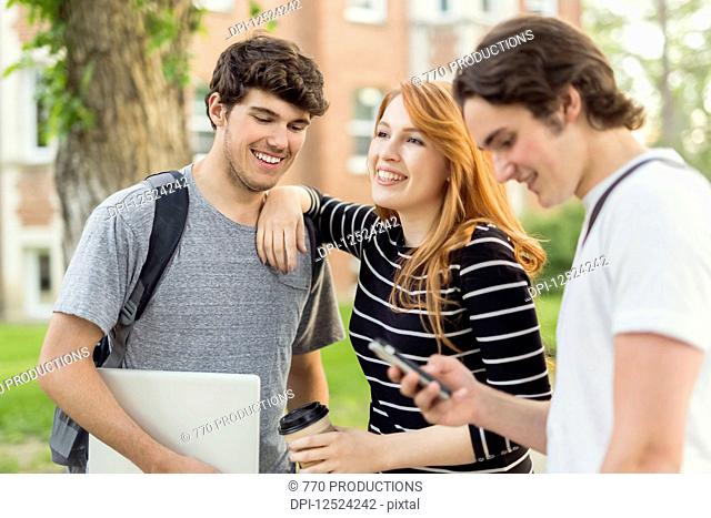 Three friends standing and talking on the university campus with their smart phone and laptop; Edmonton, Alberta, Canada
