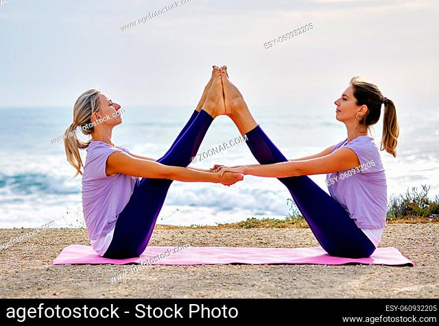 Two yogi women in activewear do yoga exercise sit on coast make wide legged buddy boat pose near sea, increase body flexibility inner balance