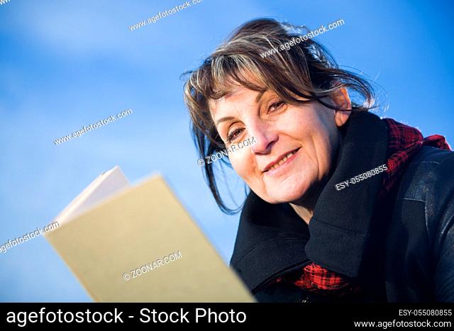 Seitliche Kopf-und-Schulter-Ansicht einer Frau gehört Alters in schwarzen Lederjacke ein Vergehen Buch haltend vor blauen Himmel in der Kamera gesehend