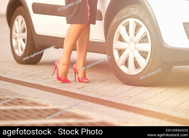 Low view of woman#39;s legs near her white car. Lady posing for photographer for fashionable mazagine. She demonstrating slim and slender legs on high heels