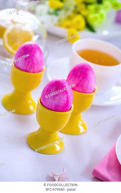 Pink Easter eggs in eggcups, cup of tea behind