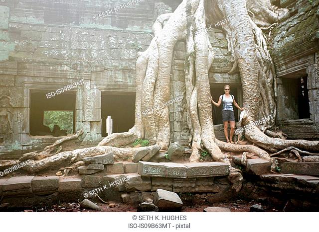 Portrait of young female tourist by tree roots at Ta Prohm temple, Angkor, Siem Reap, Cambodia