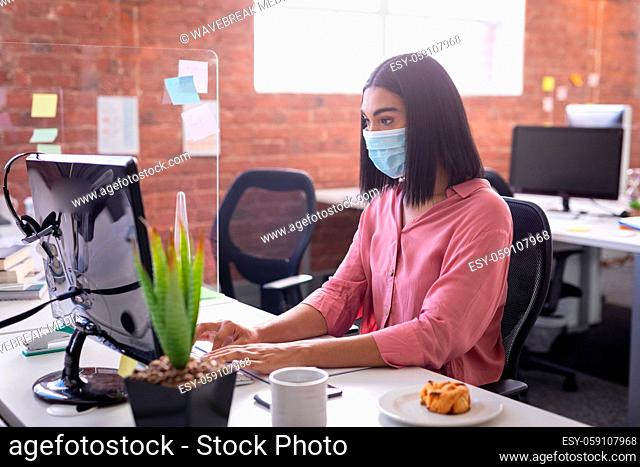 Mixed race businesswoman sitting in office in front of computer wearing face mask