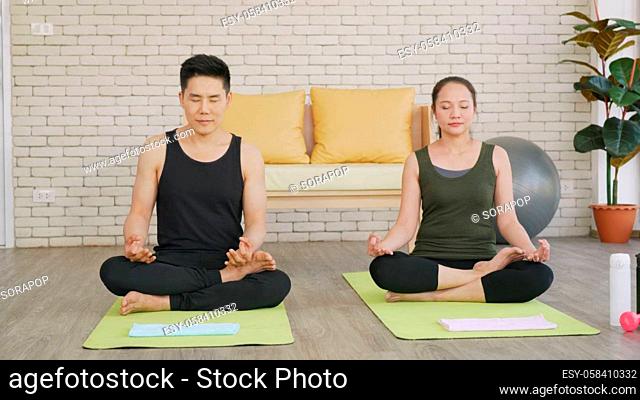Happy Asian beautiful lifestyle family couple doing YOGA sitting meditating on lotus pose workout at home together on mat with eyes closed
