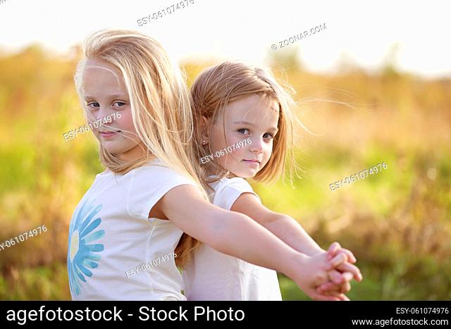 Two little girls stand with their backs to each other with their hands up in the field. Sisters