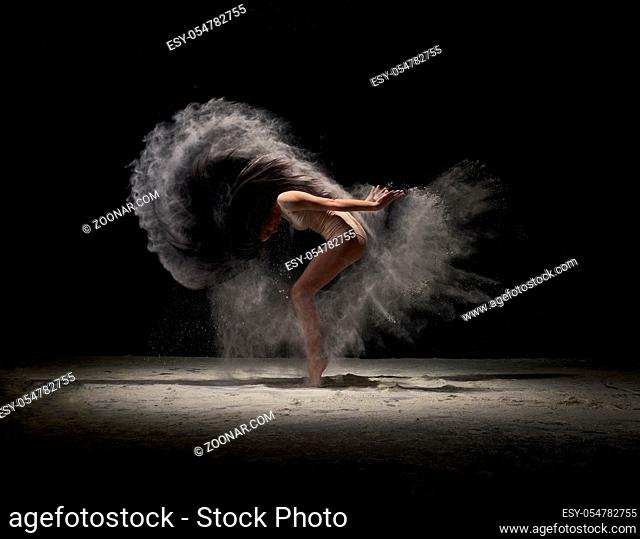 Contemporary art - full length shot of young graceful dancer at dark studio moving in cloud of powder or dust