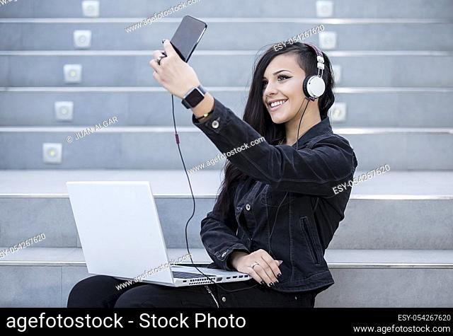 Smiling woman taking selfieï¿½while listening to music and using laptop on stairs