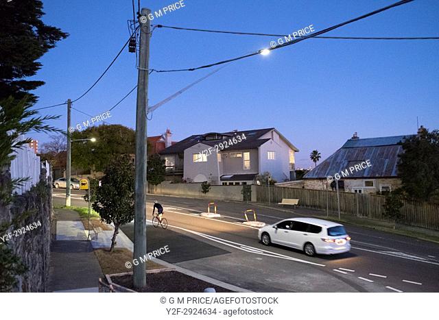 car follows cyclist on hilly suburban street at dusk, Sydney