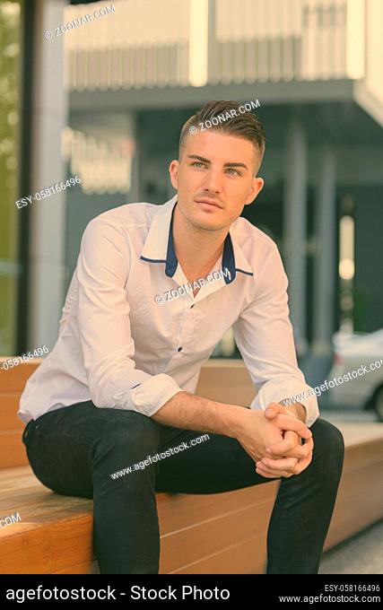 Young handsome man sitting and thinking on wooden stairs outside modern house