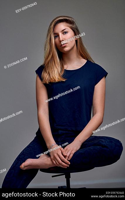 Beautiful young woman in blue t-shirt, with straight blond hair siting with one leg on chair and looking at camera. Front low angle portrait against grey...