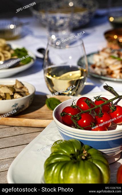 Outdoor table with tomatoes, pasta with cheese and pepper, shrimp skewers, artichokes and white wine