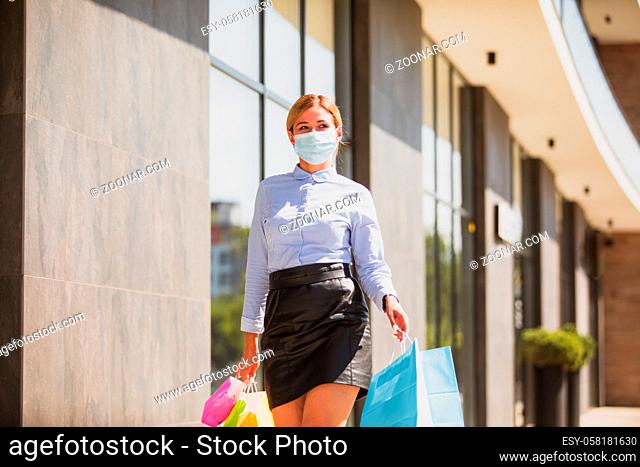 Beautiful girl in medical face mask holding shopping bags, walking down the street. Woman shopper. Coronavirus COVID-19