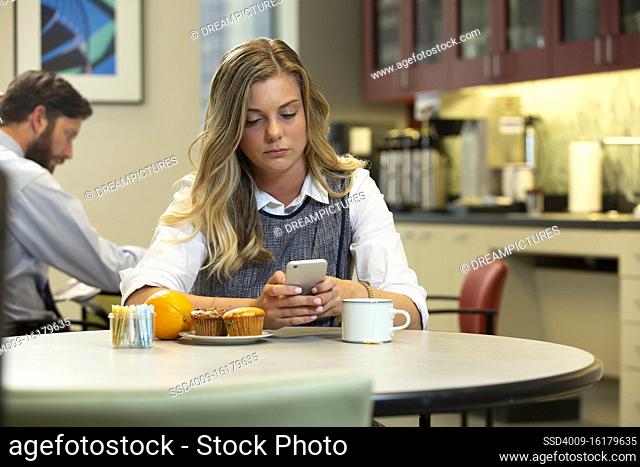 Young Caucasian woman looking at cell phone while sitting in break area of office with breakfast of an Orange, Muffins and Hot Tea