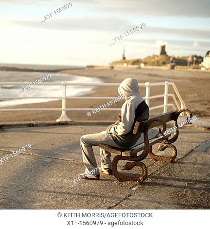 A young man wearing a hoodie sitting alone on a seaside bench, Aberystwyth Wales UK