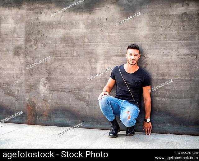 Handsome young man standing against concrete wall, looking at camera, wearing black t-shirt and jeans. Full length shot