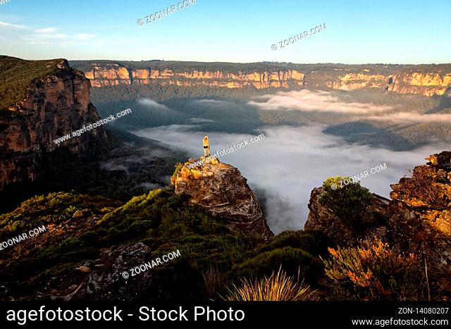 Hiker taking in magnificent views of mountains and valleys as the first light hits the sandstone escarpment cliffs and part of the valley still with a low lying...
