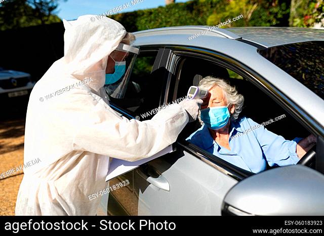 Medical worker wearing ppe suit taking temperature of senior caucasian woman sitting in car