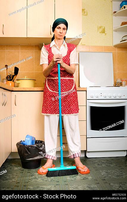 A depressed adult woman, a housewife or a maid, wearing a red apron and a green scarf on her head is resting after she has swept the kitchen with a broom