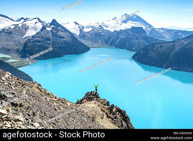 Young man standing on a rock, stretching his arms in the air, view of mountains and glacier with turquoise blue lake Garibaldi Lake, peaks Panorama Ridge